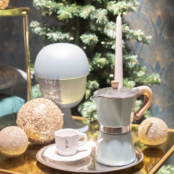 Un arrangement festif comprend une machine à expresso pour cuisinière, une tasse blanche sur une soucoupe et une bougie allumée sur un plateau. Autour, des ornements dorés scintillants et un petit sapin de Noël sont disposés devant un mur décoratif.