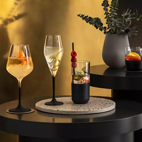 Un arrangement élégant de trois verres à cocktail élégants sur une table sombre avec un vase en céramique contenant des branches feuillues. Chaque verre contient une boisson claire et des garnitures. La scène est éclairée chaleureusement et possède un fond doré.