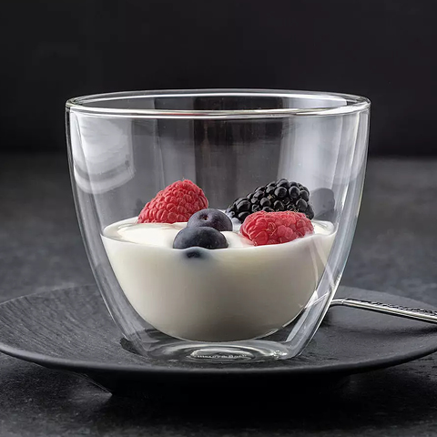 Un verre à double paroi est rempli de yaourt crémeux et garni de framboises, de mûres et de myrtilles fraîches. Le verre repose sur une soucoupe noire avec une cuillère à côté, devant un fond sombre.
