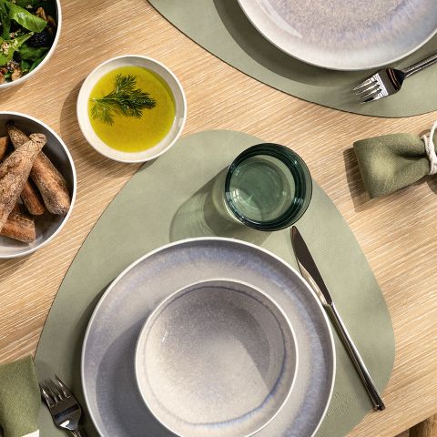 Vue de dessus d'une table à manger avec des assiettes grises, des sets de table verts, un bol de gressins, un bol de sauce verte à l'aneth et un petit verre teinté vert. Les serviettes sont soigneusement roulées et placées à côté des assiettes.