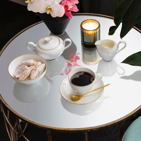 Sur une table ronde et réfléchissante se trouvent une bougie allumée, une tasse de café avec sa soucoupe et une cuillère dorée, un bol de biscuits saupoudrés de sucre glace, un sucrier, un pot à crème et un vase d'orchidées roses et blanches.
