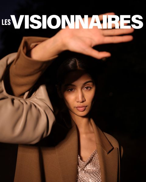 Une personne portant un manteau marron lève une main pour cacher partiellement son visage. Elle se tient devant un fond sombre. En haut de l'image, les mots « Les Visionnaires » sont écrits en lettres blanches grasses.