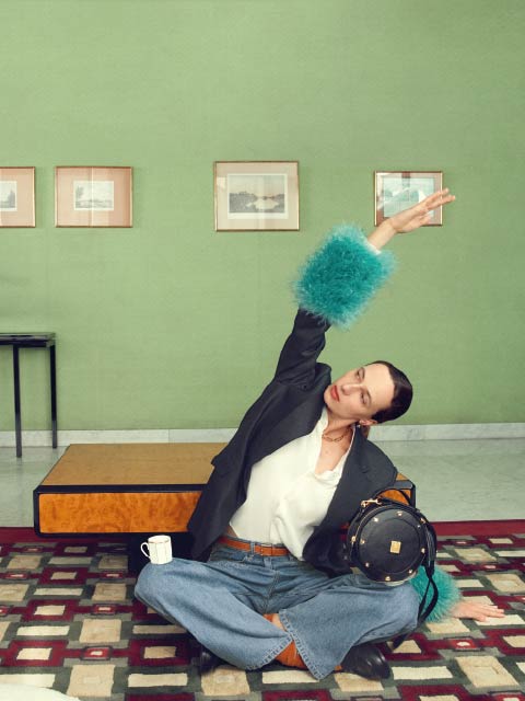 Une personne en jean et blazer avec des poignets bleus et pelucheux pose le bras levé dans une pièce élégante aux murs verts, des œuvres d'art encadrées et un tapis à motifs.