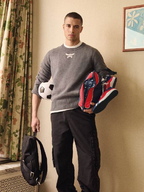 Un jeune homme en pull gris tient un sac à dos noir et des baskets à la main, un ballon de football sous le bras, et se tient à l'intérieur près de rideaux fleuris.