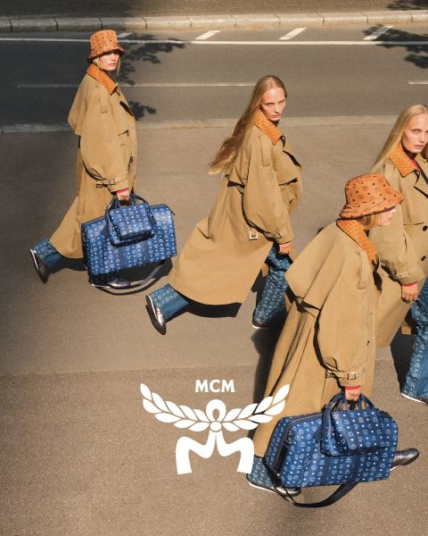 Trois femmes portant des manteaux marron clair et des chapeaux orange traversent une rue, portant des sacs MCM bleus avec un logo MCM blanc au premier plan. L'image a un effet miroir qui donne l'impression que la scène est dupliquée.