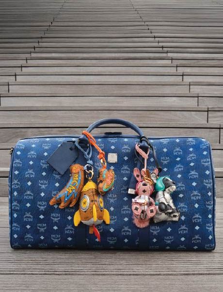 Un sac de créateur MCM bleu avec un motif de logo blanc est posé sur des marches en bois ornées de porte-clés colorés représentant des animaux et des objets, notamment une fusée, un lapin et un ours.