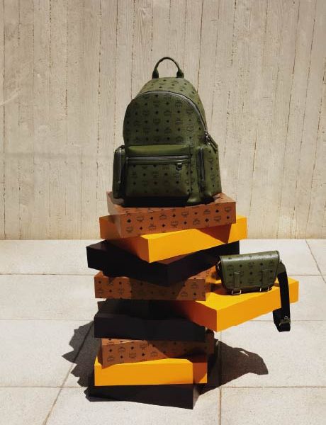 Un sac à dos de créateur vert et un sac à bandoulière assorti sont posés sur une pile de boîtes orange et marron devant un mur texturé beige et un sol carrelé.