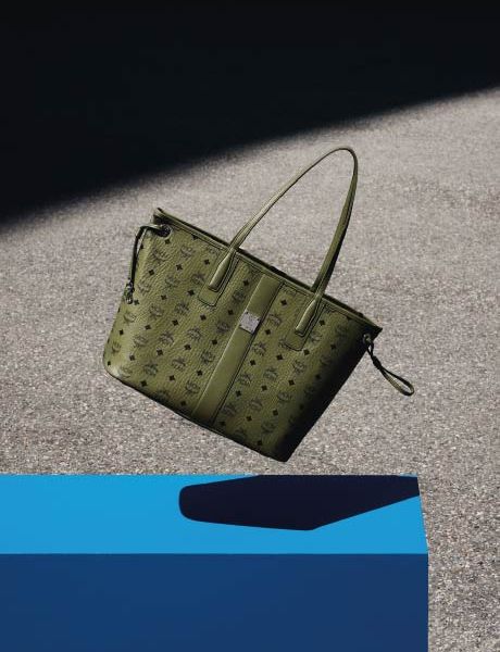 Un sac à main de créateur vert avec un motif monogramme discret repose sur une surface grise, avec un objet géométrique bleu au premier plan et une ombre portée à l'arrière-plan.
