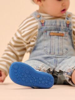 Un jeune enfant en chemise rayée et salopette en jean est assis avec une jambe à l’extérieur et montre la semelle bleue texturée de sa chaussure.