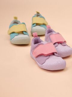 Deux paires de chaussures pour tout-petits sur fond beige : une paire est lilas clair avec des brides roses, l'autre bleu clair avec des brides jaunes.