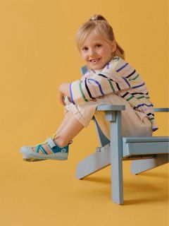 Enfant souriant portant une chemise à rayures et un pantalon clair, assis de profil sur une chaise en bois bleue devant un fond jaune.