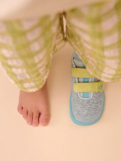 Un pied d’enfant nu à côté d’une chaussure de sport avec des bandes jaunes, tous deux visibles sous un pantalon à carreaux vert clair sur un sol beige.