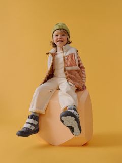 Un enfant souriant avec un bonnet vert, une chemise rayée et un gilet est assis sur un bloc couleur pêche devant un fond jaune, portant des bottes et un pantalon clair.