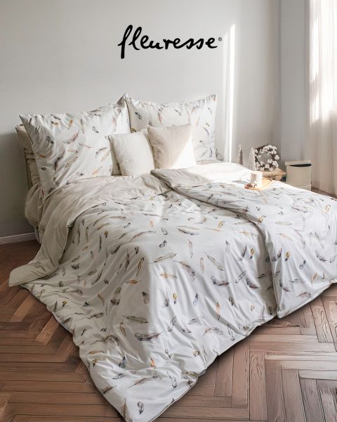 Une chambre confortable avec un lit bien fait, recouvert de draps blancs à motifs d'oiseaux. Deux coussins décoratifs sont posés sur la tête de lit. La lumière du soleil filtre à travers des rideaux transparents, projetant une douce lueur sur le plancher en bois clair.