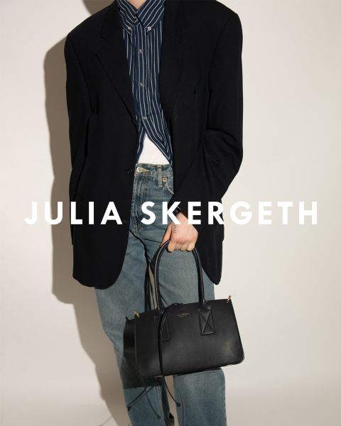 Personne portant un blazer noir sur une chemise rayée et un jean bleu, tenant un sac à main noir. Le texte "JULIA SKERGETH" est clairement visible superposé à l'image.