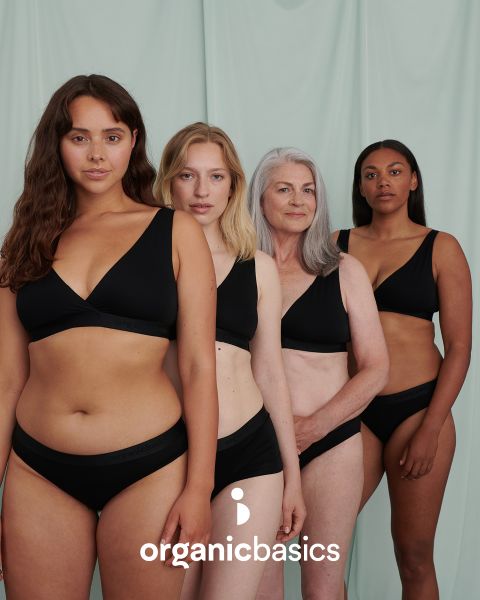 Quatre femmes d'âges et de morphologies différents se tiennent avec assurance devant un fond vert clair, vêtues de sous-vêtements noirs assortis. Le texte "organicbasics" apparaît en bas de l'image.