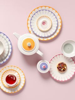 Assiettes colorées à motifs floraux, une théière, une tasse de thé avec citron et une pâtisserie, arrangées sur un fond rose.