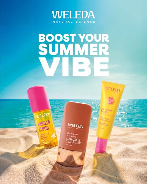 Trois produits de soin Weleda dans le sable avec la mer et le ciel bleu en arrière-plan. Texte : « Boost Your Summer Vibe ». Scène de plage lumineuse et estivale.