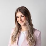 Une femme aux longs cheveux châtain clair sourit doucement et porte un blazer rose clair sur un haut blanc devant un fond blanc uni.