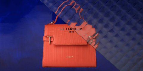 Un élégant sac à main orange Le Tanneur est présenté devant un fond texturé bleu vif. Le sac a un design chic avec une fermeture à rabat et le nom de la marque « Le Tanneur 1898 » en relief.