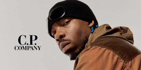 Homme portant une veste marron, un bonnet noir avec des lunettes de protection et des perles dans les cheveux. Il regarde l'appareil photo avec une expression sérieuse. Sur le côté gauche, devant un fond uni, se trouve le texte "C.P. Company".