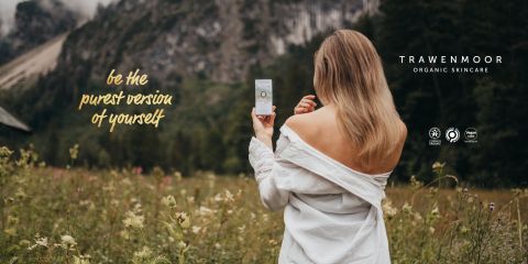 Une femme aux longs cheveux blonds, le visage tourné, se tient dans un champ et tient un produit de soin de la peau à la main. Elle porte un haut à épaules dénudées. Le texte est : "Sois la version la plus pure de toi-même." À l'arrière-plan, on aperçoit des montagnes et de l'herbe. L'écran affiche "TRAWENMOOR ORGANIC SKINCARE".