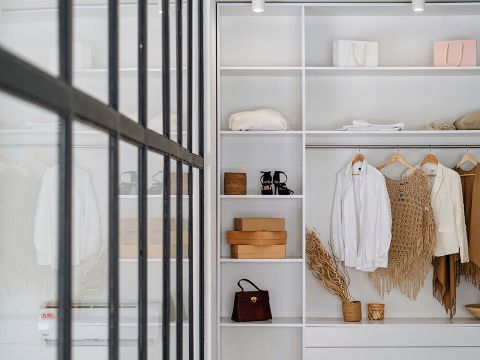 Une garde-robe bien organisée avec des étagères et des cintres blancs. Elle contient des vêtements, des sacs à main et des boîtes marron. Une paroi de verre reflète partiellement l'intérieur et contribue au design moderne.
