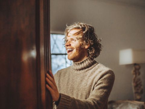 Une personne aux cheveux bouclés et à la barbe porte un pull à col roulé beige et douillet. Elle sourit avec une expression joyeuse et regarde sur le côté. La pièce est éclairée chaleureusement, avec une fenêtre et une lampe en arrière-plan.