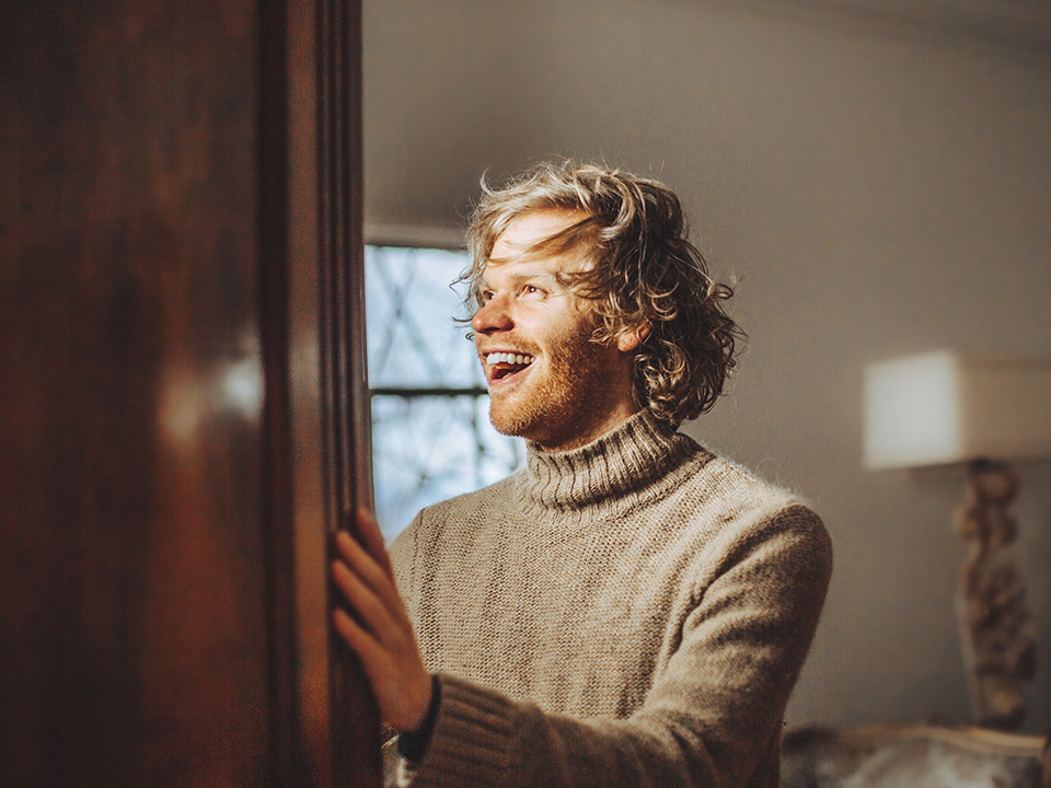 Une personne aux cheveux bouclés et à la barbe porte un pull à col roulé beige et douillet. Elle sourit avec une expression joyeuse et regarde sur le côté. La pièce est éclairée chaleureusement, avec une fenêtre et une lampe en arrière-plan.