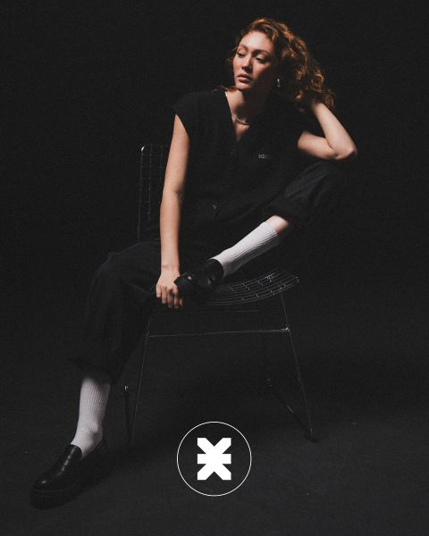 Une personne aux cheveux bouclés est assise sur une chaise en fil de fer dans une pièce sombre, vêtue de vêtements noirs, de chaussettes blanches et de chaussures basses noires. Une jambe est pliée, un coude repose sur le genou, et sur le sol se trouve un logo circulaire avec un "X".