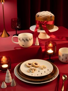 Une table dressée pour une fête avec un grand gâteau, entouré d'un ruban rouge et saupoudré de sucre glace, sur un présentoir. Il y a des biscuits sur des assiettes, des gobelets à thème, des bougies, des décorations de Noël rouges et blanches et un fond rouge foncé.