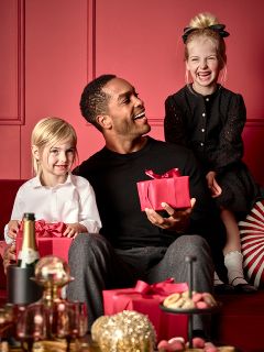 Un homme est assis sur un canapé rouge avec une boîte cadeau rouge et sourit à deux jeunes enfants. Un enfant, vêtu de blanc, tient un cadeau rouge similaire, tandis que l'autre, vêtu de noir, est assis sur le dossier du canapé. Au premier plan, il y a une table avec des friandises et une bouteille.