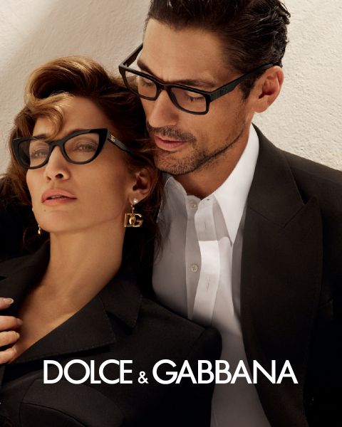 Une femme et un homme portant d'élégantes lunettes sont appuyés l'un contre l'autre. Elle porte des boucles d'oreilles DG et une tenue noire, il porte un costume noir avec une chemise blanche. Le logo de Dolce & Gabbana est visible en bas.