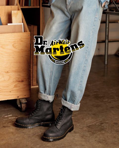Une personne portant un jean bleu clair et des bottes à lacets noires Dr. Martens se tient à l'intérieur sur un sol en béton. Le logo « Dr. Martens AirWair » est clairement visible au-dessus de l'image. À l'arrière-plan, on aperçoit une étagère en bois et un pied de chaise en métal.
