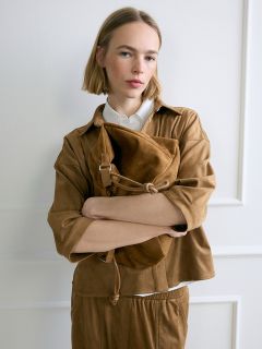 Une femme avec un carré blond, vêtue d'une tenue marron, se tient les bras croisés devant un mur blanc, tenant un grand sac en daim marron.