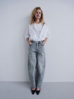 Une femme en chemise blanche et jean gris ample se tient devant un mur blanc, les mains dans les poches, portant des escarpins noirs et regardant l'appareil photo.