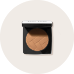 Ein Kompaktpuder mit Bräunungspuder in einem quadratischen schwarzen Etui mit geöffnetem Deckel, der einen warmen Braunton mit strukturierter Oberfläche offenbart. Der Deckel ist weiß mit schwarzem Text. Das Produkt ist auf einem schlichten, hellbeigen Hintergrund zu sehen.