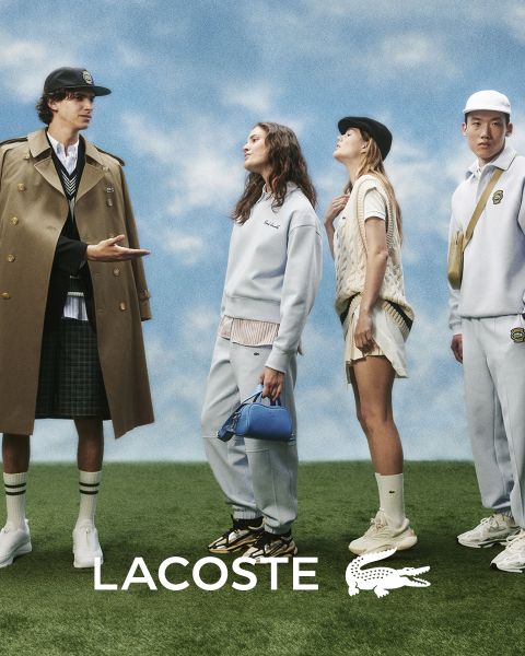 Quatre mannequins posent en extérieur, vêtus de tenues Lacoste, devant un ciel bleu. De gauche à droite : une personne en trench-coat, une personne en pull et pantalon, une personne en gilet et jupe, et une personne en survêtement. Le logo Lacoste se trouve en bas.
