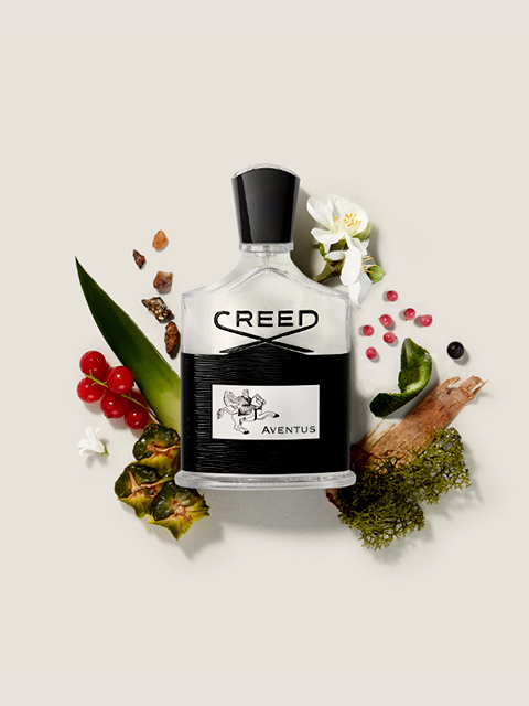 Une bouteille en verre de parfum Creed Aventus, entourée de ses ingrédients : baies rouges, fleurs blanches, ananas, mousse et quelques épices sombres. L'arrière-plan est d'un beige doux.