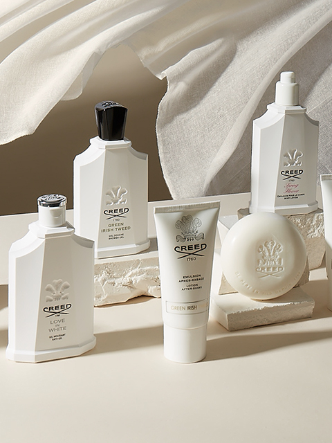 Une présentation luxueuse des produits de soin de la peau et des parfums Creed, comprenant des flacons, un tube et un pain de savon. Tous les articles sont élégamment disposés sur une surface aux tons neutres avec un tissu doux et fluide en arrière-plan.