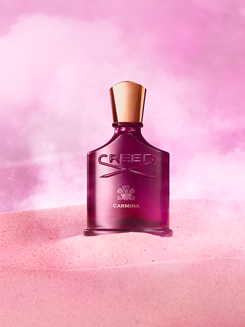 Un flacon rose de parfum Creed Carmina est posé devant un fond rose brumeux, reposant sur une surface qui ressemble à du sable rose. Le flacon a un bouchon doré et affiche le nom de la marque et le logo en relief.