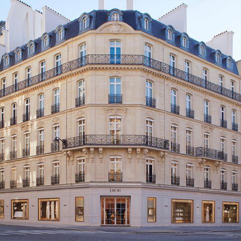 Élégant bâtiment d'angle à l'architecture classique, avec plusieurs étages, des fenêtres cintrées et des balcons en fer forgé ouvragés. Au rez-de-chaussée se trouve une boutique Dior avec de grandes vitrines. La lumière naturelle vive rehausse la façade beige.