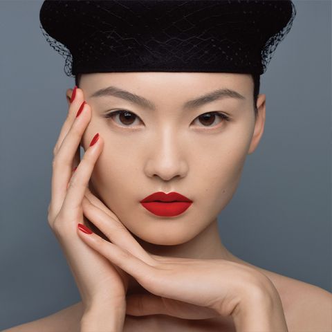 Une personne portant un rouge à lèvres et un vernis à ongles rouge vif pose devant un fond gris. Elle porte un chapeau noir avec une structure en résille et ses mains sont élégamment placées autour de son visage, soulignant le maquillage éclatant.
