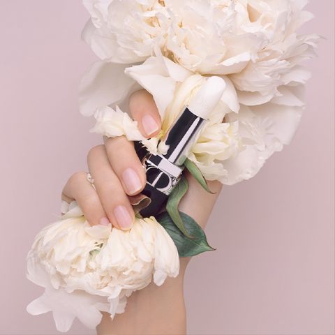 Une main aux ongles manucurés tient un rouge à lèvres blanc, entourée de grandes fleurs de pivoines blanches, créant une atmosphère douce et élégante. L'arrière-plan est rose clair, ce qui complète l'esthétique délicate.