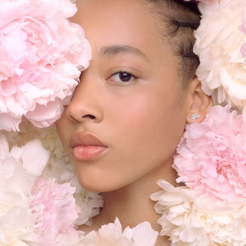 Une femme est partiellement entourée de grandes pivoines roses et blanches. Son visage est doucement éclairé, révélant un maquillage naturel et des lèvres brillantes. Elle porte une petite boucle d'oreille élégante à son oreille visible. Le ton général de l'image est doux et serein.