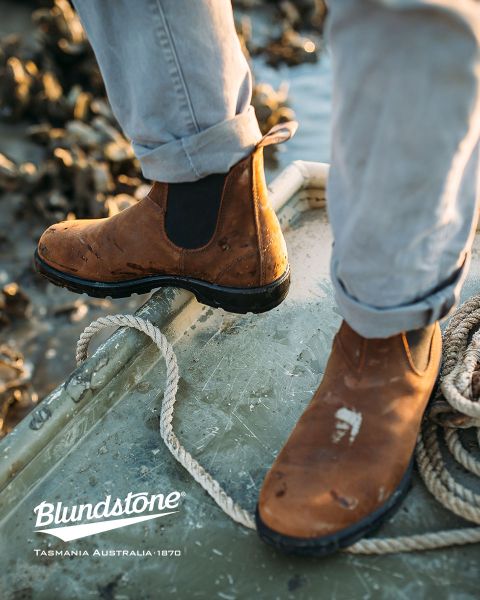 Une personne portant des bottes Blundstone marron usées se tient sur un bateau délabré près de cordages, avec de l'eau et des rochers flous en arrière-plan.