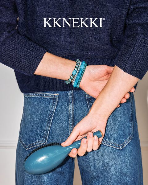 Une personne en pull bleu marine et jean tient une brosse à cheveux bleue derrière son dos et porte des bracelets de perles turquoise. Au-dessus, le logo "KKNEKKI".