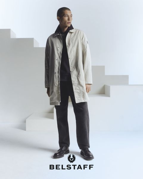 Une personne portant un manteau clair sur un pantalon et des chaussures sombres se tient devant un fond minimaliste et géométrique. Le logo « Belstaff » est visible en bas de l'image.