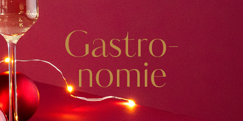 Un verre de vin blanc et une guirlande lumineuse sont placés à côté d'une décoration de Noël rouge devant un fond rouge vif. Le mot « Gastronomie » est écrit en lettres dorées.