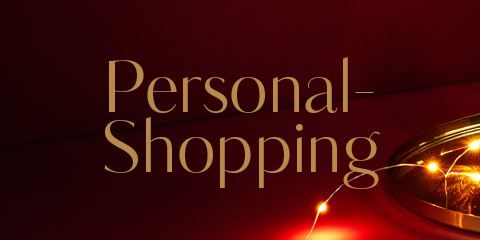 L'image montre l'inscription « Personal-Shopping » en élégante écriture dorée sur un fond rouge foncé avec des lumières décoratives discrètes.